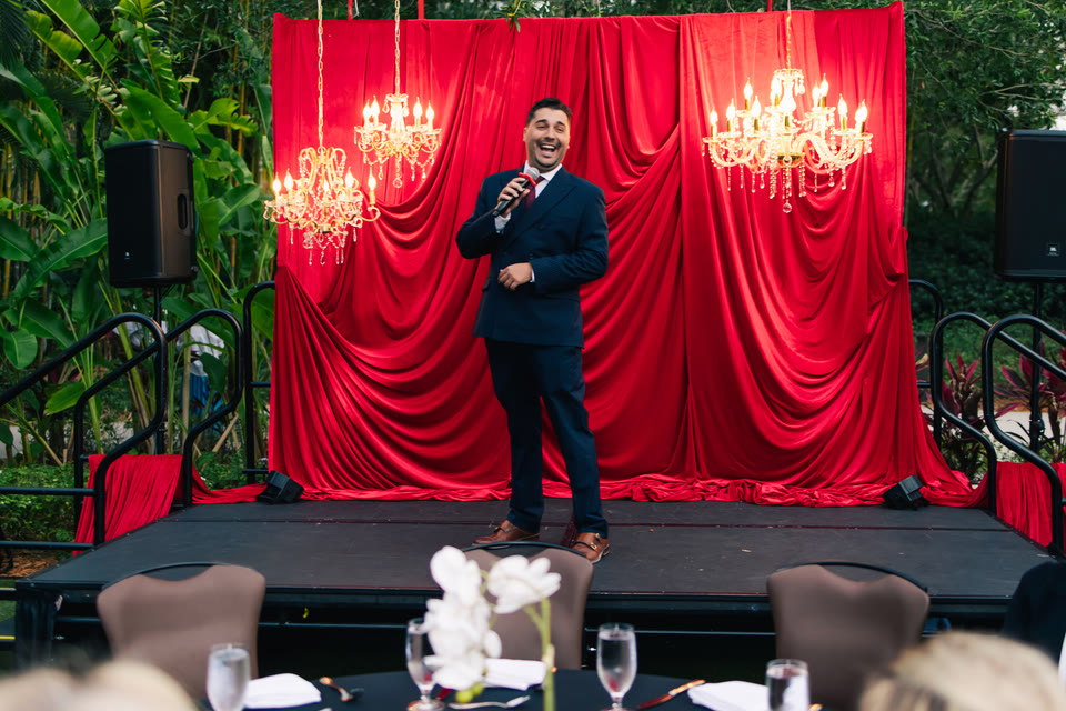 Performer on stage under chandeliers, with a deep-red curtain backdrop.